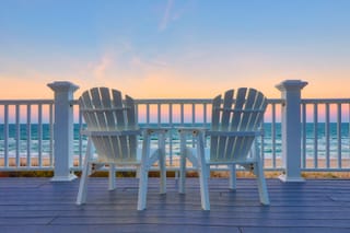 Image of two empty adirondack chairs on a deck overlooking a beach at sunset