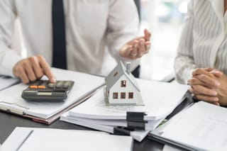 Image of two people using a calculator with a small house sitting on papers on a desk