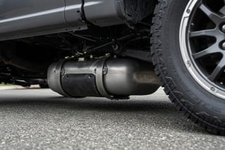 Fuel tank held by two metal straps beneath a pickup truck parked on asphalt