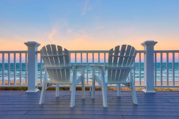 Image of two empty adirondack chairs on a deck overlooking a beach at sunset