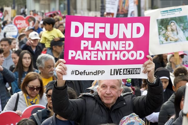 Photo of a protest with an attendee holding a sign stating 'Defund Planned Parenthood' 