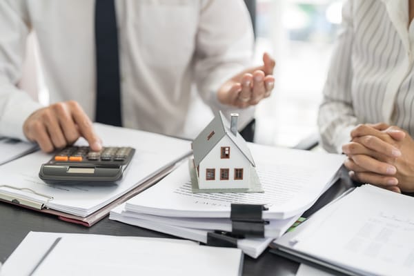 Image of two people using a calculator with a small house sitting on papers on a desk