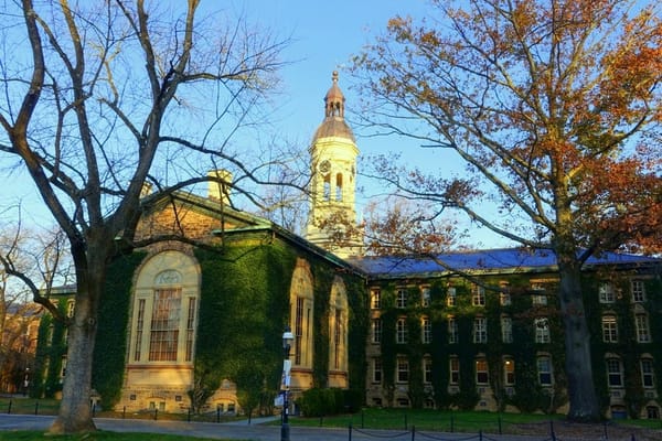 Ivy-covered Nassau Hall at Princeton University, with its cupola and tall arched windows, framed by autumn trees under a blue sky.